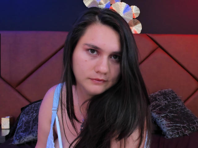 AvienLume has long black hair, a light blue top, and a metallic headpiece. She has a serious expression with subtle makeup and a small pimple on her right cheek.