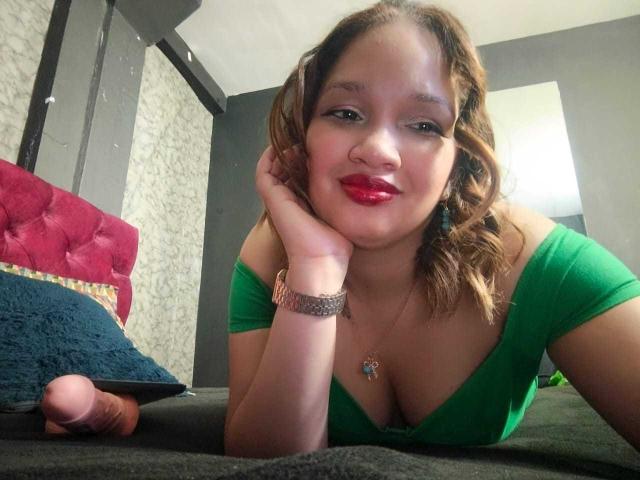 BiancaMorettii, com batom vermelho, blusa verde e cabelo castanho ondulado, deitada na cama, com a cabeça apoiada na mão, mostrando um pênis flácido e não circuncidado à esquerda..