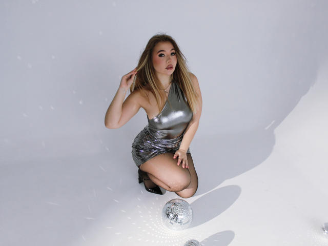 Stacy Yellig, con lunghi capelli biondi, indossa un abito argentato aderente, è inginocchiata su uno sfondo bianco, con una mano tra i capelli, indossa tacchi neri e davanti a lei c'è una sfera da discoteca argentata..