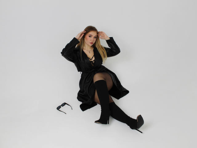 Stacy Yellig está sentada no chão branco, vestindo um vestido preto, meias finas e botas que chegam até o joelho, com as mãos no cabelo loiro, e óculos de grau pretos ao lado dela, mostrando suas nádegas..
