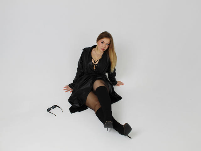 Stacy Yellig assise sur un sol blanc, portant une robe noire à lacets sur le devant, des bas noirs jusqu'aux genoux, des talons hauts noirs, et des lunettes sur le sol, cheveux blonds, sexy, regardant la caméra..