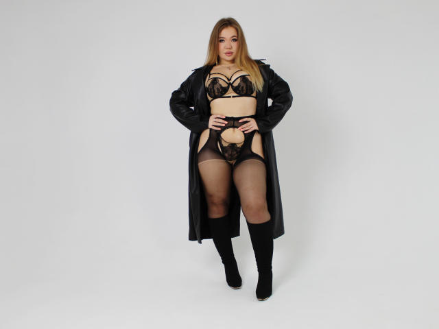 Stacy Yellig está em pé, vestindo lingerie de renda preta, um casaco comprido, meias finas e botas. Ela tem cabelos loiros longos e um corpo curvilíneo e sexy, com seios proeminentes e um bumbum pequeno..