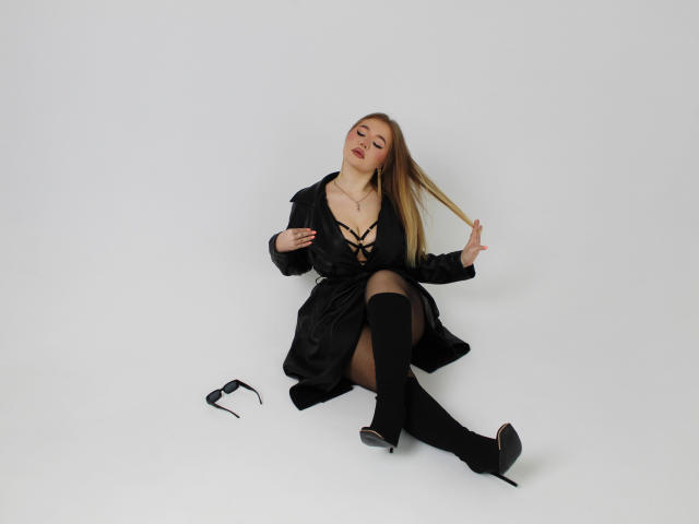 StacyYellig is sitting on the floor with black boots and a black dress, her tits visible through the lace-up front, with glasses next to her.