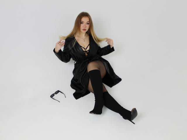 StacyYellig with blonde hair, black dress, lace-up cleavage, black stockings, and knee-high boots, sitting on white floor with black sunglasses beside her.