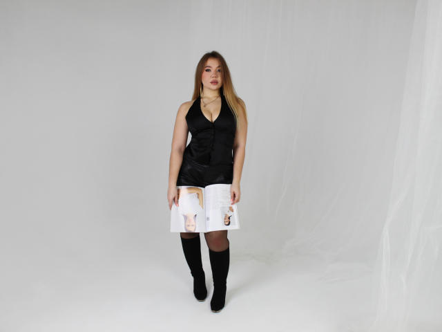 Stacy Yellig staat daar, gekleed in een zwarte top met halter, een witte rok met bedrukte afbeeldingen en zwarte kniehoge laarzen. Ze heeft blond haar, een lichte huid en een subtiele glimlach..
