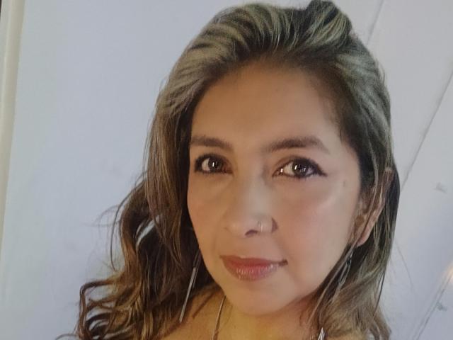 MichelleDumont has brown wavy hair, brown eyes, and a subtle smile with shiny pink lips. She's wearing small earrings and has a light skin tone. Her tits are not visible.