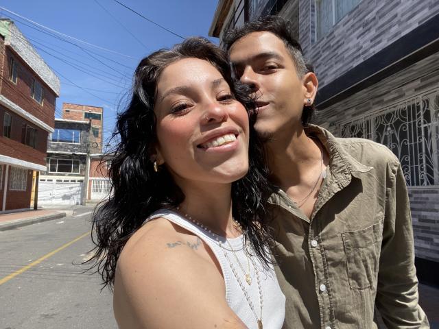 Yara Leon, con cabello oscuro y una camiseta blanca, sonríe y tiene un tatuaje en el hombro. Está al lado de un hombre con cabello corto y una camisa verde..