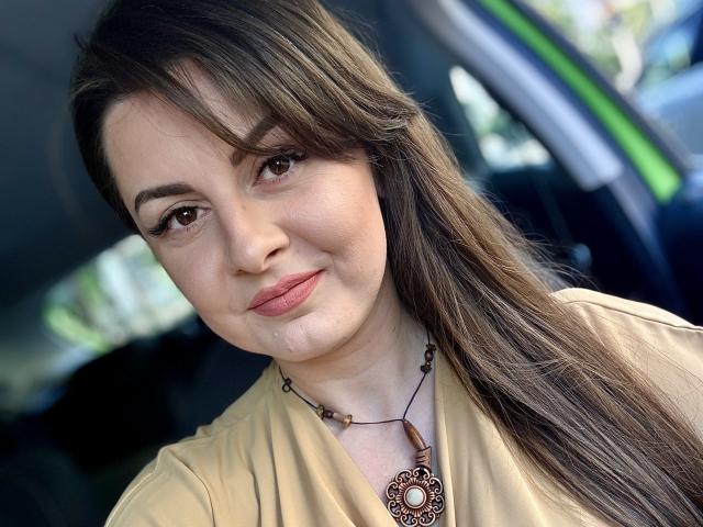 HelleBoobs has long brown hair, brown eyes, light skin, and a slight smile. She's wearing a beige shirt with a brown necklace, sitting in a car.