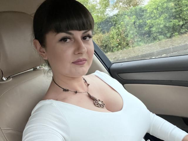 HelleBoobs, com cabelo castanho escuro preso em um coque, vestindo uma blusa branca e um colar, sentada em um carro, mostrando seus seios, olhando para a câmera, com um leve sorriso..