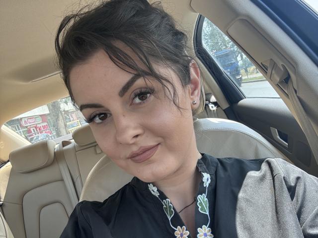 HelleBoobs, con el cabello oscuro recogido en un moño despeinado, vestida con una camisa negra con estampado floral, está sentada en un coche, sonriendo suavemente, mostrando pequeños senos, y lleva un piercing en la nariz..