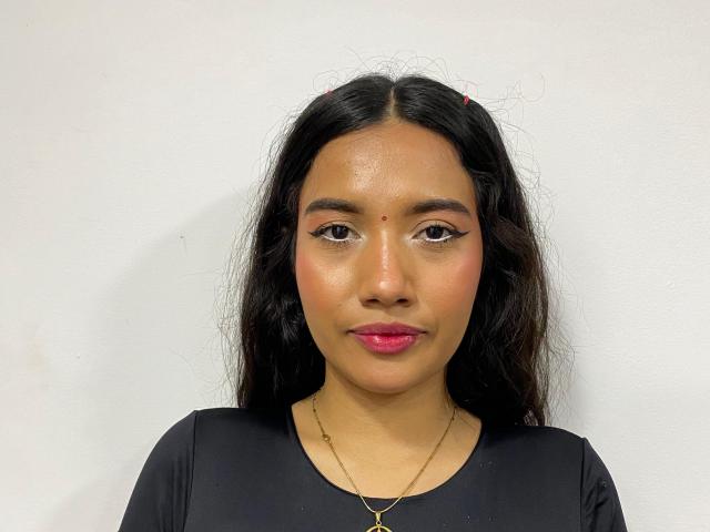 AnneLover heeft lang zwart haar, donkere wenkbrauwen, roze lippenstift en een kleine moedervlek op haar neus. Ze draagt een zwarte top en een gouden ketting..