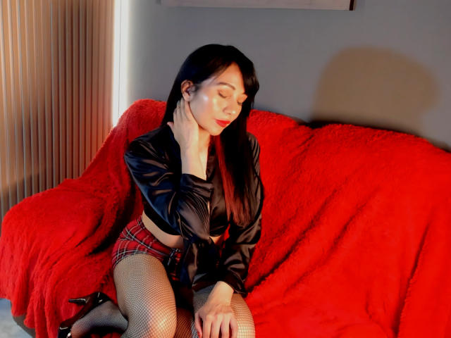 AkaneNoir69 with black hair, red lips, black satin top, plaid skirt, fishnet stockings, and black heels, sitting on a red couch, eyes closed, hand touching neck.