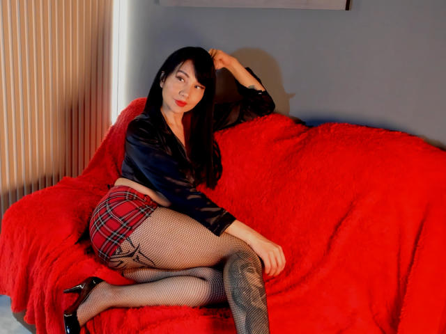 AkaneNoir69 with black hair, red plaid skirt, fishnet stockings, black heels, and black velvet top, laying on a red fluffy couch, hand in hair, looking at camera.