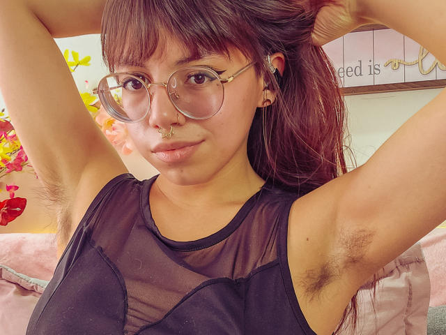 NaturalMariaHun with armpit hair, wearing clear glasses, a black sheer top, and a nose ring, looking at the camera with arms raised. She has brown hair and a cute face.