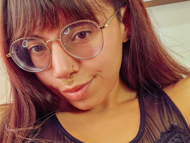 NaturalMariaHun with brown eyes, wearing gold-rimmed glasses, a nose ring, and a black sheer top, has a slight smile and a cute, soft look.