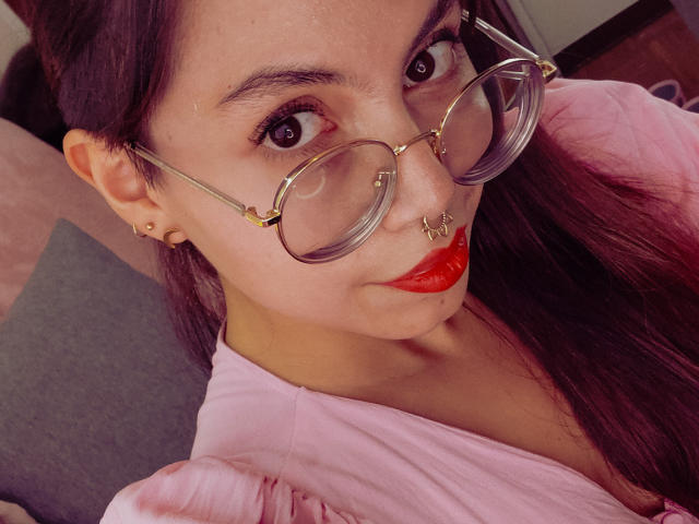 NaturalMariaHun with red lipstick, nose ring, septum ring, glasses, and pink shirt, has long purple hair and a cute face, looking at the camera with big brown eyes.
