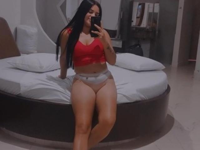 GiovannaSafad with long black hair, red bra, and grey panties, sitting on a bed, holding a phone, showing her curvy ass and thick thighs in a mirror selfie.