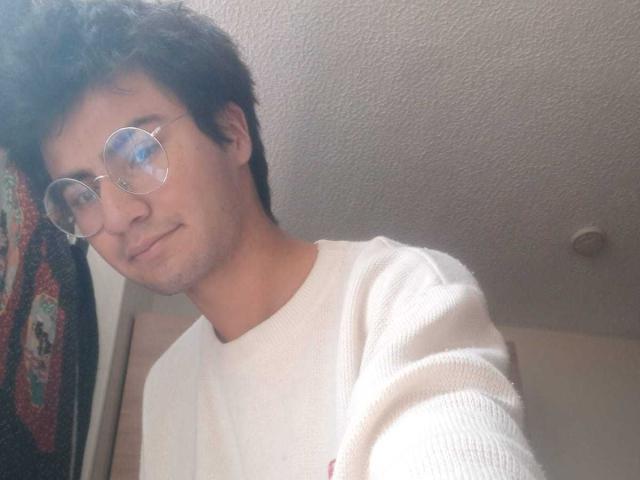 ElioVega con gli occhiali, capelli corti e scuri, pelle chiara, maglione bianco, leggero sorriso, texture del soffitto, parziale decorazione colorata a parete, selfie casual..