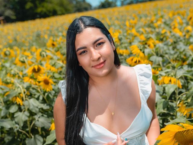 AuroraSxx with long black hair, white dress, and cleavage, holding a sunflower, smiling in a sunflower field, wearing a gold necklace. Her tits are small and cute.