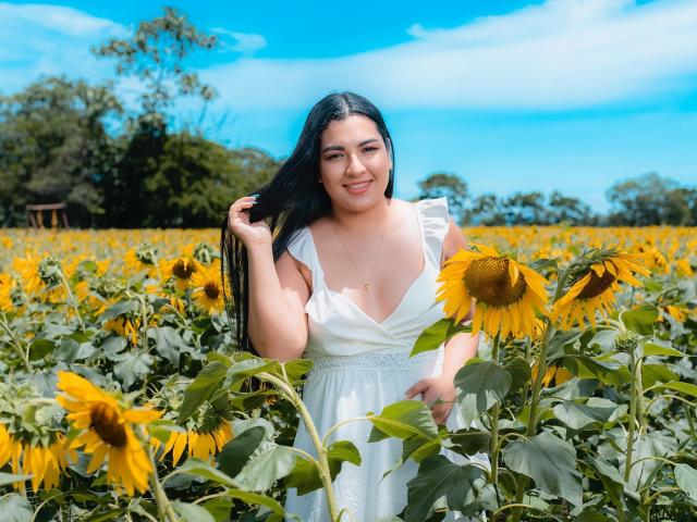 AuroraSxx with black hair, white dress, and cleavage, smiling in a sunflower field, holding her hair, blue sky, and green leaves, sunny day, standing among tall sunflowers.