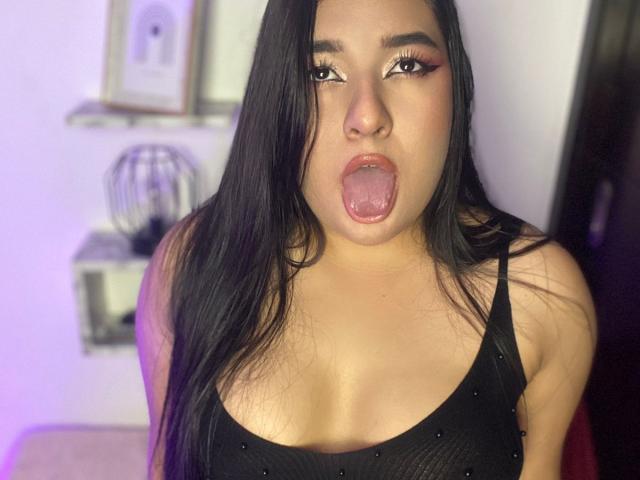 AuroraSxx with long black hair, wearing a black tank top, has her tongue out, showing off her puffy, glossy lips and looking directly at the camera.