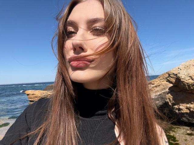 SensualMila ha lunghi capelli castani, un maglione nero a collo alto e labbra rosa carnose, con un cielo blu limpido e una costa rocciosa sullo sfondo. Il suo viso è leggermente scompigliato dal vento..