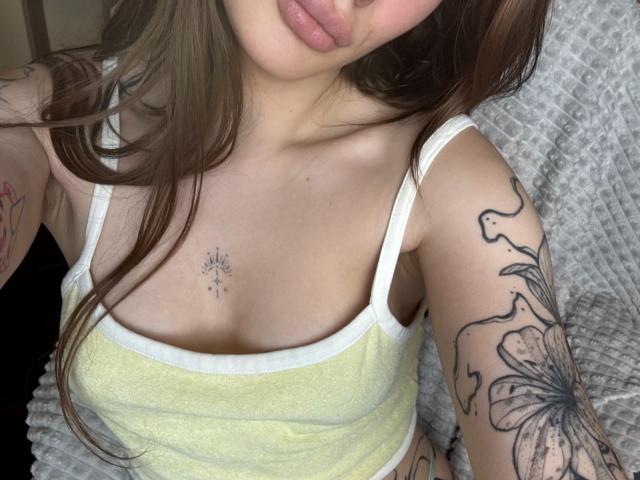 SensualMila with light brown hair, wearing a yellow tank top, showing tattoos on her arm and chest, and her small tits. Her pouty lips are slightly visible.