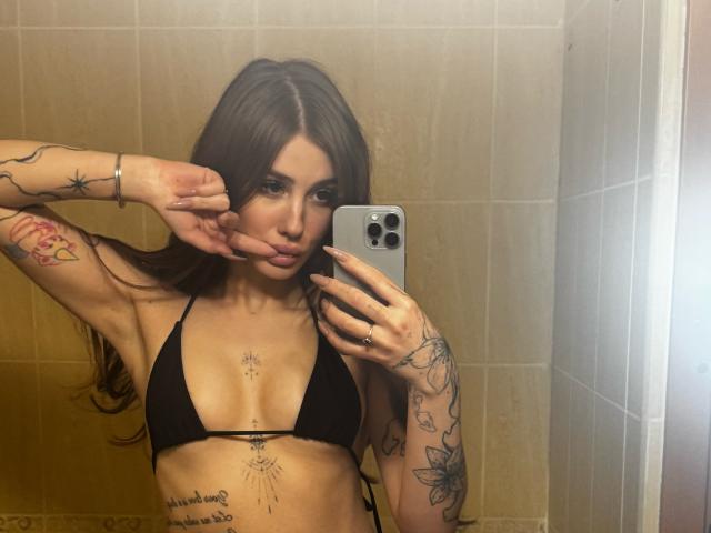 SensualMila with long brown hair, topless, showing tattoos, small black bra, and red plaid skirt, holding phone to mirror, in bathroom, side profile, showing her tits.