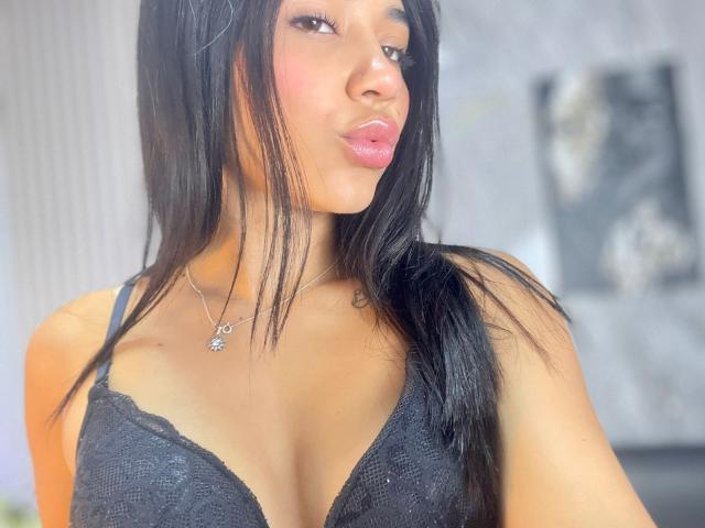 NahomyCarther with long black hair, hoop earrings, and a black off-shoulder top with lace-up front, showing small tits, smiling softly, standing in a room.
