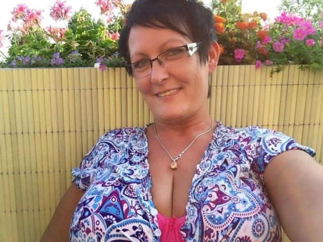 Sexy milf met kort zwart haar, gekleed in een witte tanktop, staat op een houten zeilboot, met in haar handen het roer, met een touw en zeil op de achtergrond..