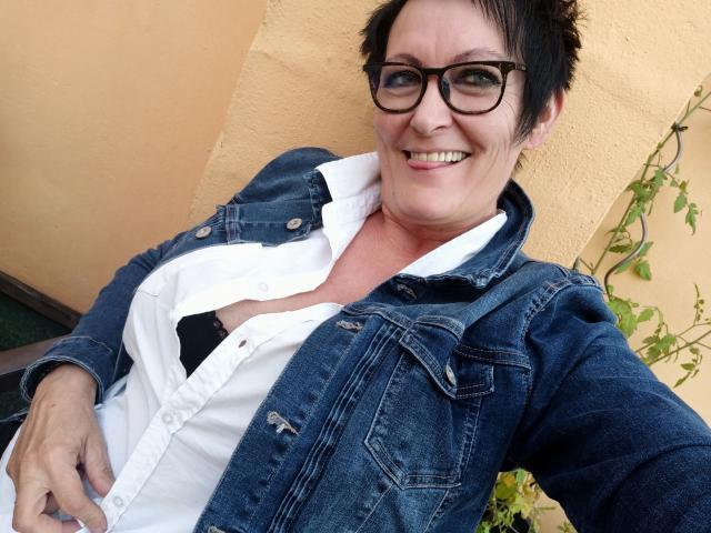 Una MILF attraente con capelli corti e scuri, occhiali e un sorriso, indossa una maglietta blu e viola con un motivo che mette in risalto il suo seno generoso, e una collana d'oro, ed è in piedi all'aperto..
