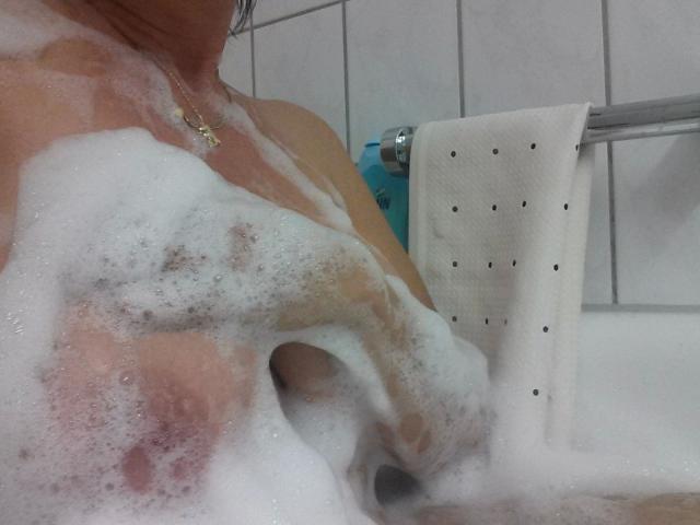 Uma mulher atraente, com cabelo preto curto, óculos e um sorriso, vestindo uma jaqueta jeans azul por cima de uma camisa branca de botões, um sutiã preto visível e uma tatuagem na mão direita..