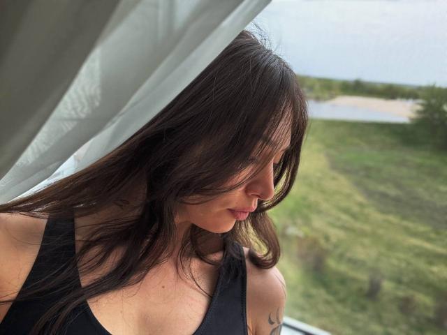 MoniFoxy met golvend bruin haar, bruine ogen en donkere lipstick, die haar arm omhoog steekt en haar gezicht en bovenlichaam laat zien, gekleed in een grijze top, op een zonnige selfie..
