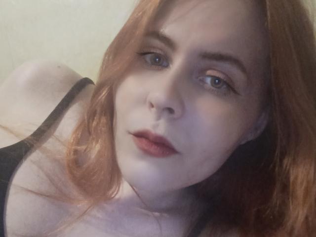 MistressKat con seni di media grandezza, una maglietta rossa, senza biancheria intima, un tatuaggio a forma di stella sul pube, capelli neri, un orologio al polso, seduta sul letto, gambe divaricate, con un'espressione sorpresa..