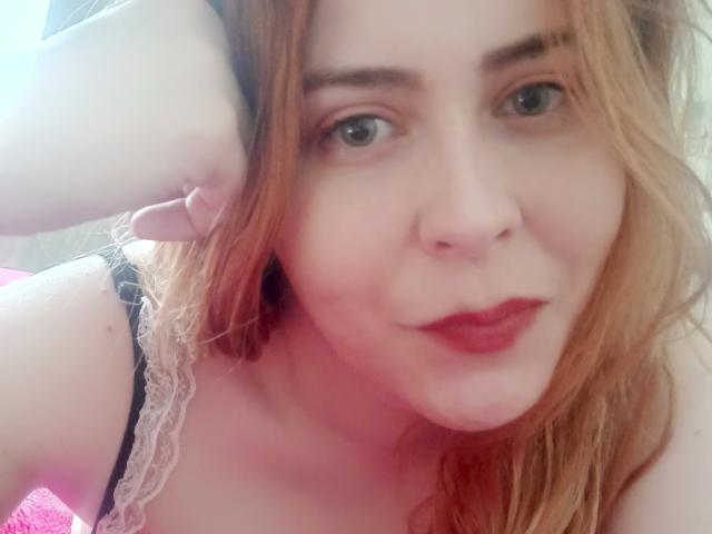 FannyNorah, con cabello rojo, piel pálida y lápiz labial rosa, mirando hacia la cámara. Lleva puesto un top negro con encaje, que deja ver sus pechos. Fondo rojo..