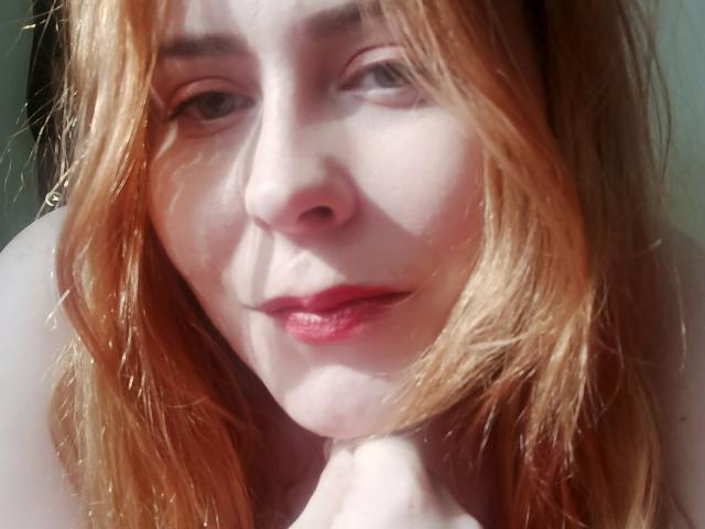 FannyNorah, com batom vermelho, pele clara e cabelo loiro, vestindo um sutiã de renda preta com detalhes brancos, mostrando seus seios, olhando para a câmera com uma expressão de beicinho..