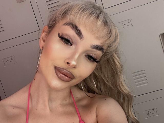 PollyDance tiene el cabello rubio recogido en una coleta, lleva una blusa rosa, tiene pestañas largas, cejas marcadas y labios rosados brillantes. Está mirando hacia arriba con una ligera sonrisa..