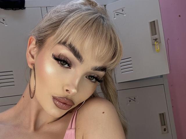 PollyDance a les cheveux blonds, des sourcils épais, de longs cils, des lèvres brunes brillantes, un haut rose à encolure américaine et de grandes boucles d'oreilles. Elle regarde l'appareil photo avec un léger sourire..