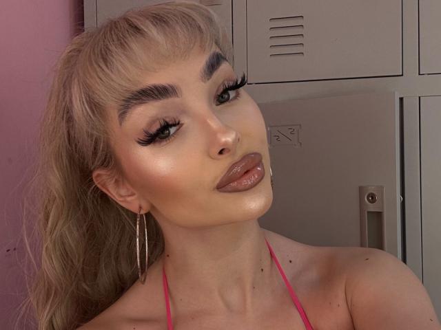 PollyDance a les cheveux blonds, les yeux verts et des cils épais, elle porte de la lingerie rose, des boucles d'oreilles cerceaux et un rouge à lèvres rose brillant, avec une expression sexy et séduisante et une peau éclatante..