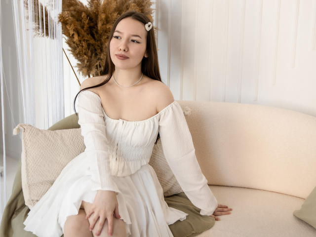 DanielleSweetty with long brown hair, white off-shoulder top, and subtle smile, holding a sheer curtain, looking at camera, showing her tits, soft light, delicate necklace.