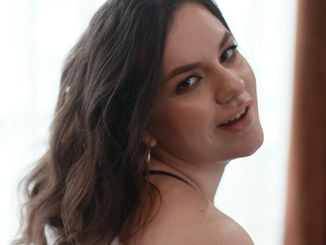 CharlotteCrofort with long black hair, smiling with pink lipstick, wearing pink flower earrings, and a silver necklace, has a cute nose ring and a shiny face.