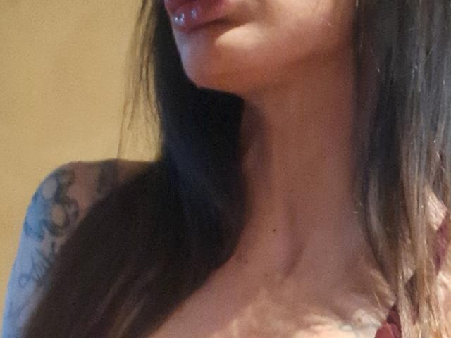 LadyMilena69's upper chest and face, showing glossy lips, a tattoo on her right shoulder, and brown hair, with her tits slightly visible under a patterned top.