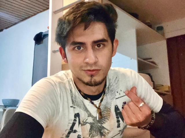 SamCooperX, com cabelo castanho curto, sobrancelhas escuras e barba, vestindo uma camisa branca com uma estampa de pássaro, apontando para o seu anel com um sorriso discreto..