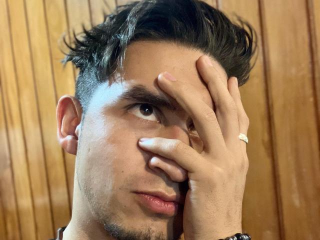SamCooperX with short, dark, messy hair, covering one eye with a hand, wearing a ring on his finger, showing a slightly confused or tired expression.