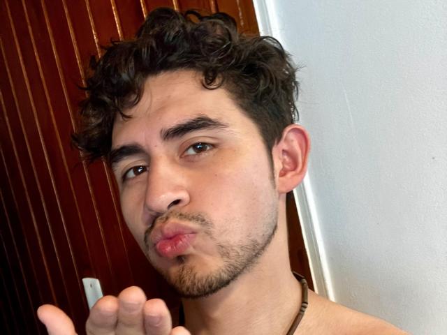 SamCooperX met krullend donker haar, bruine ogen, lichte huid en een baard, een selfie met opgetrokken lippen, die een grappig gezicht trekt, met een vinger omhoog, en een bruine ketting om..