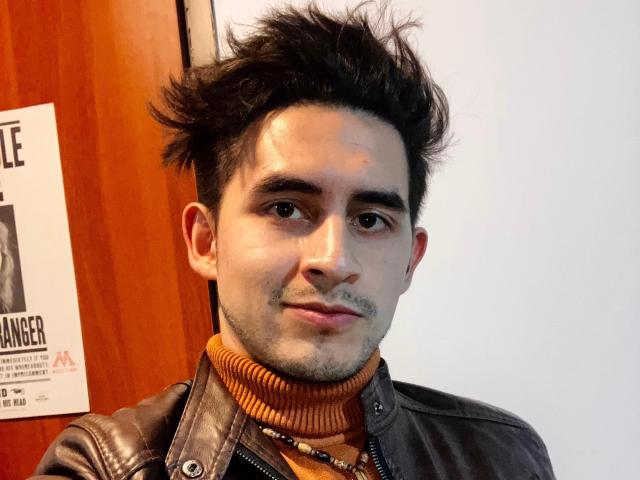SamCooperX with spiky dark hair, brown leather jacket, orange turtleneck, and slight stubble, smiling at the camera, showcasing a small tattoo under his right eye.