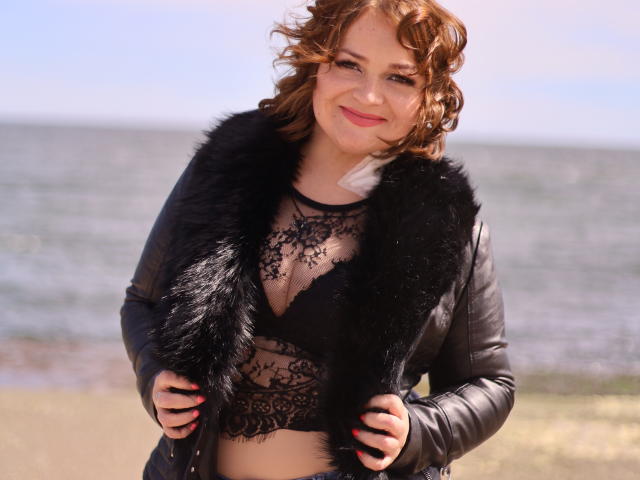 Helena Bitton, con i capelli rossi ricci, indossa un reggiseno di pizzo nero e una giacca di pelle con fodera in pelliccia, è in piedi vicino all'oceano, sorride, mostra il seno, tiene la giacca, ha le unghie rosse..