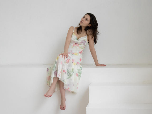 LounaLove sits barefoot on a white step, wearing a floral dress, her tits slightly visible, looking up with a dreamy expression, her ass resting on the step.