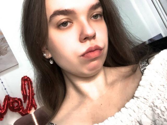 KatrinePearl has long brown hair, light skin, and small tits, wearing a white textured sweater, with a serious expression, and pearl earrings in a selfie.