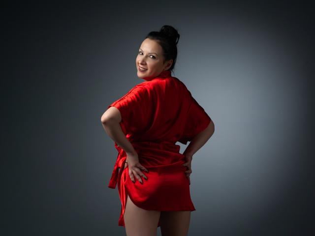 ElizabethSo, with black hair in a bun, wearing a red dress, standing with hands on hips, smiling over her shoulder, showing her ass. Dark gray background.